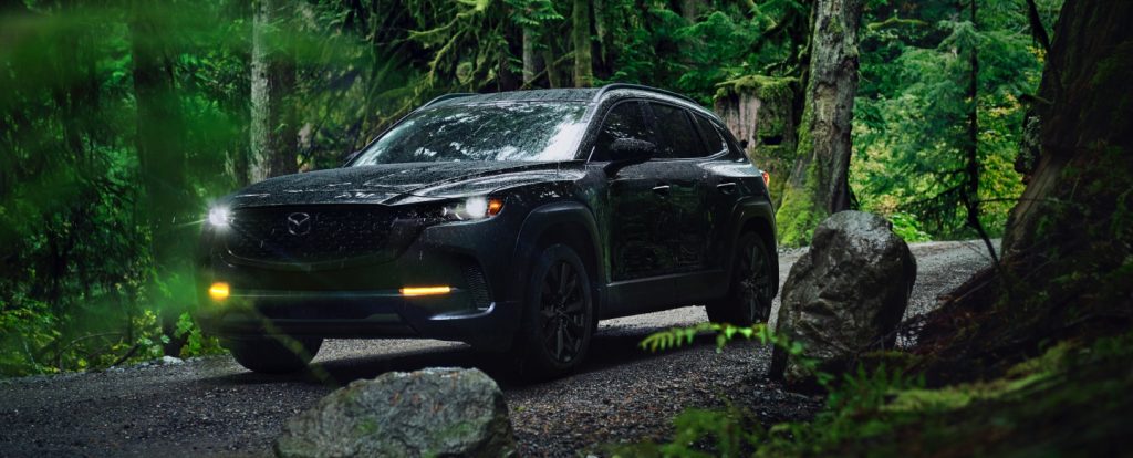 Black Mazda CX-50 driving on a gravel path through a lush forest.
